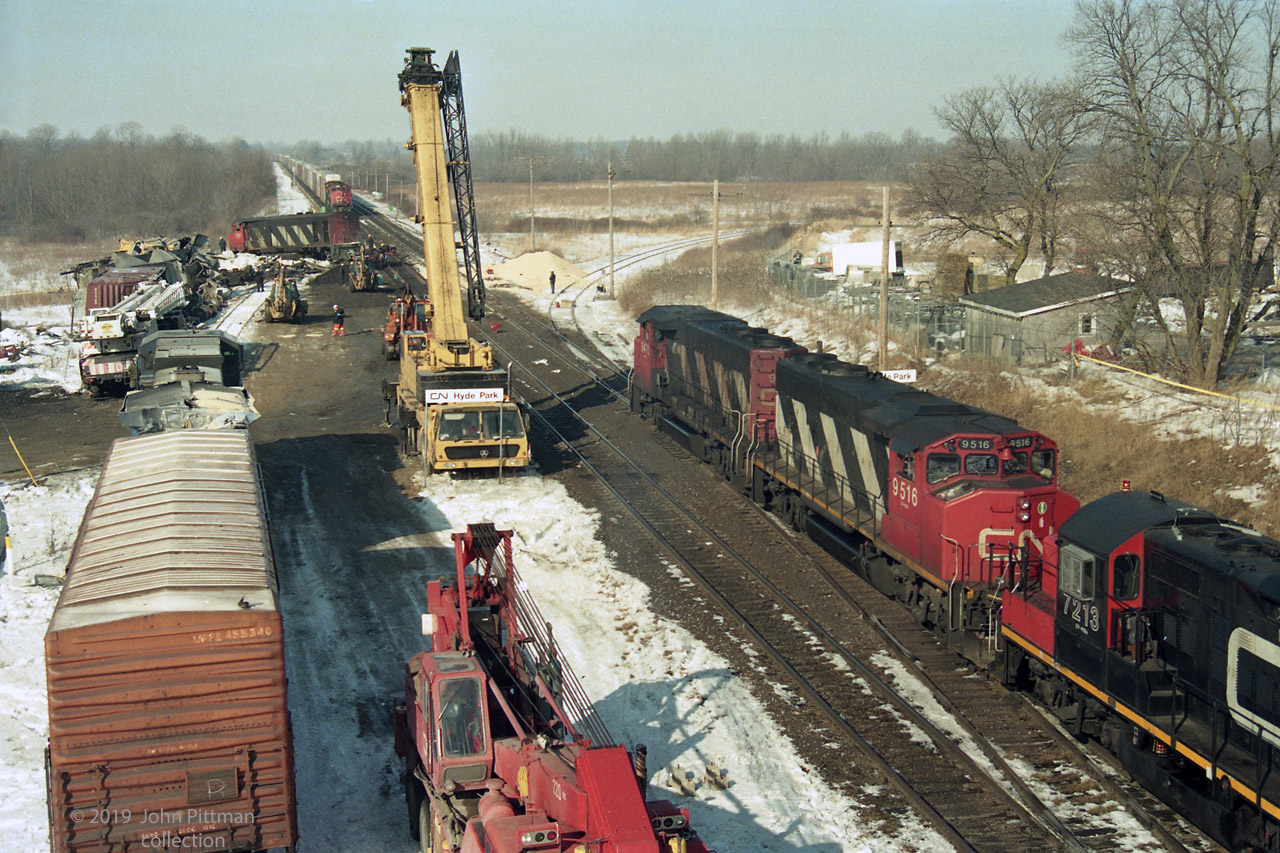 Submitted on the 24th anniversary of the Hyde Park train crash.  I recommend the TSB Report about this mishap - http://www.bst-tsb.gc.ca/eng/rapports-reports/rail/1995/r95s0021/r95s0021.asp 
 This is a non-telephoto view of the accident scene, seen from the Hyde Park Road overpass. It could be the day of the crash, or a day or more later. A westbound train is proceeding forward from the bottom right on the now-clear north track, with lead unit CN9470, CN9516, CN7213. 
Accident cleanup must have been aided by the level ground, available siding track, and good road access at the western fringe of London ON.  Three cranes are on scene and two backhoes are visible, with men at work on the damaged south track.  Derailed HR616 CN 2105, lead unit on train 272, remains where it landed - it originally faced eastward toward the vantage point. Mangled railcars have been hauled away from the tracks. A pile of light coloured material, between the spur track and the north main, is thought to be from a hopper car destroyed in the crash. Train 272's damaged trailing unit LMSX 723 had been re-railed and moved to the siding  (still tail-end first), along with a boxcar damaged in the crash. The facing train of autoracks looks like the main part of train 272, in which case rescue locomotives are on the front of it.  
You can click on "Photographer" for a couple of other views, including prior-to-cleanup image 36332.